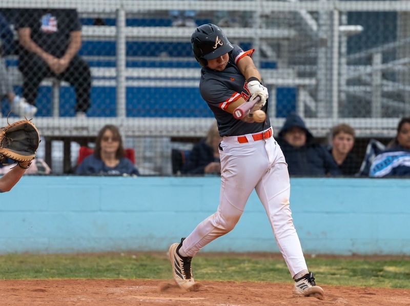 A teen boy in a dark grey and orange baseball uniform swings as the bat connects with the ball.