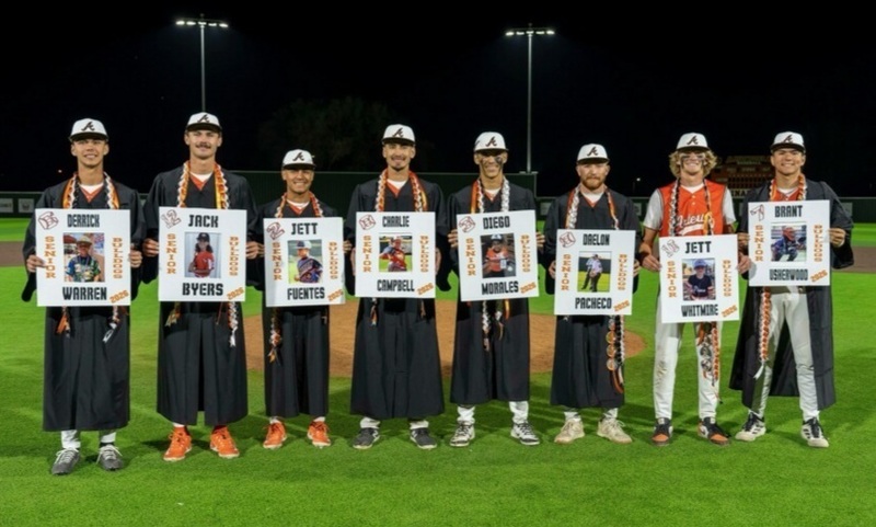 Eight teen boys in black graduation gowns stand in a row and hold signs featuring photos of themselves.