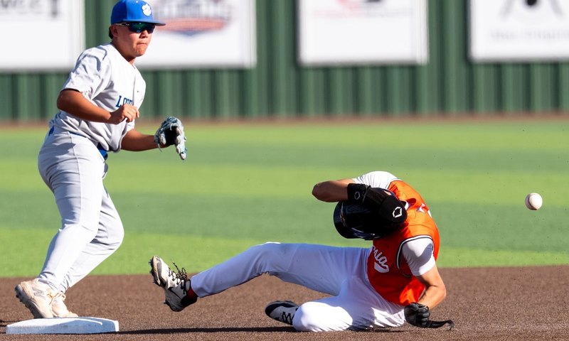 A teen boy in an orange and white baseball uniform ducks as he slides into second. An opponent in grey and blue can be seen looking at the baseball as it bounces toward the outfield.