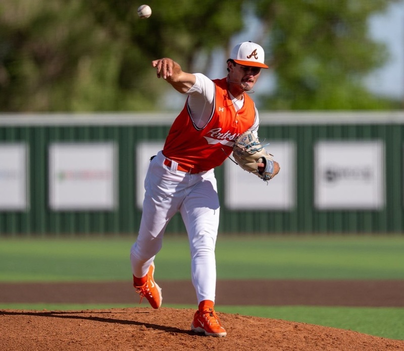 A teen boy in an orange and white baseball uniform lunges forward as he delivers a pitch from the mound.