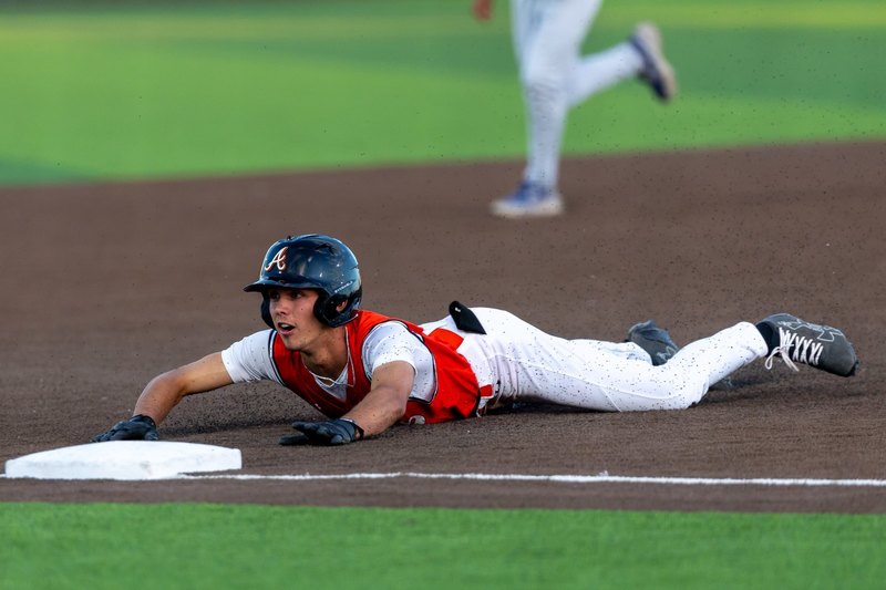 A teen boy in an orange and white baseball uniform looks up as he slides head first into third base.