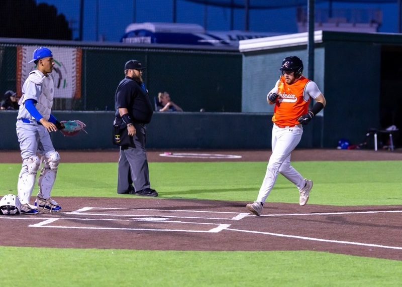 A teen boy in an orange and white baseball uniform prepares to step on home plate as an opponent in grey and blue and an umpire look on.