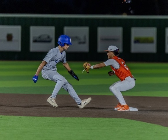 A teen boy in an orange and white baseball uniform stands on second base and prepares to tag an opponent in blue and grey who is attempting to return to the base.
