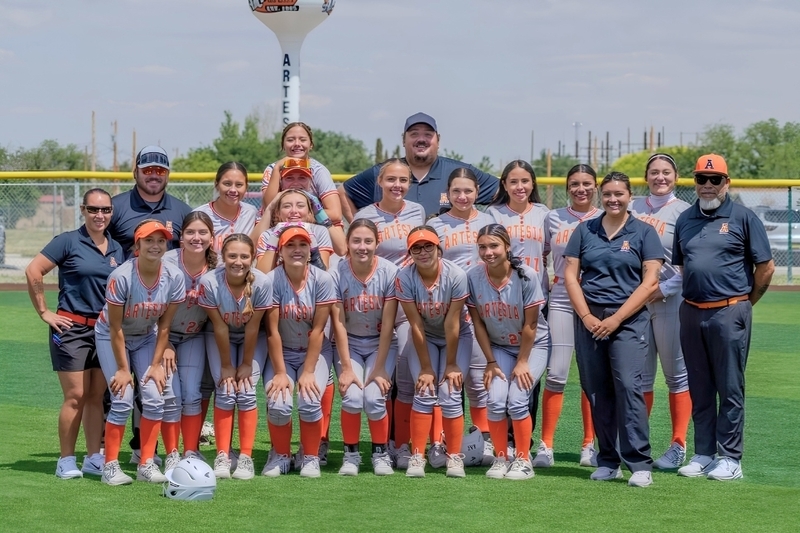A high school softball team in grey and orange uniforms poses for a photo in an outfield.