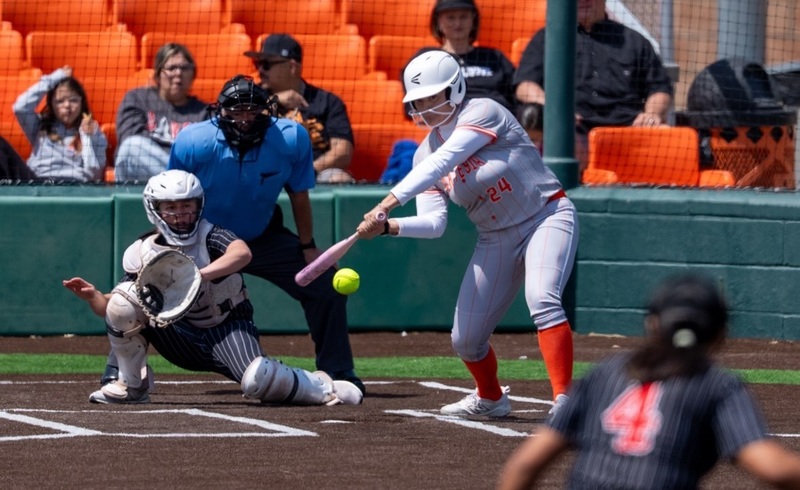 A teen girl in a grey and orange softball uniform leans toward home plate as she prepares to put the bat on the ball. Also visible are the opposing catcher and an umpire.