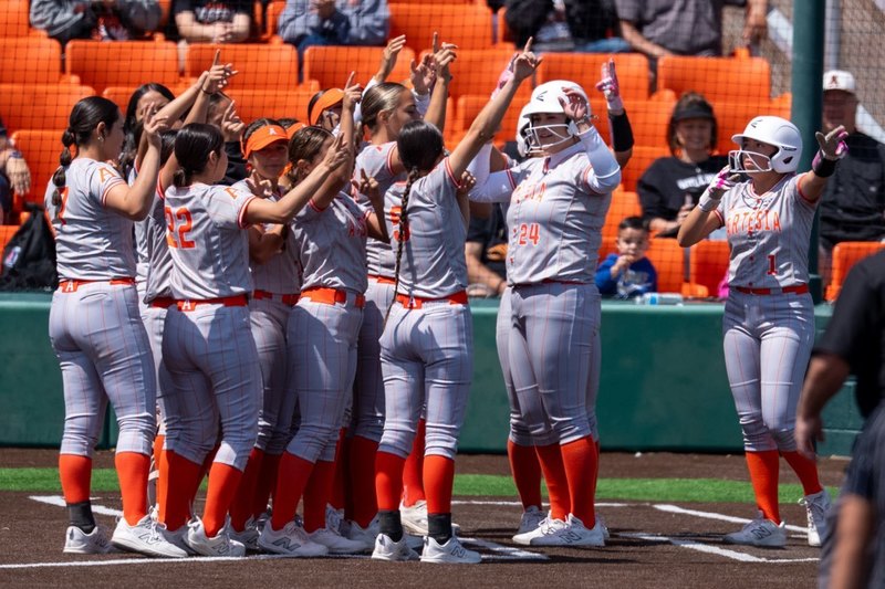 A teen girl in a grey and orange softball uniform steps on home plate and is greeted by a large group of teammates.