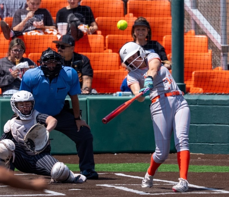 A teen girl in a grey and orange softball uniform completes her swing as the ball is seen flying into the air. Also visible are the opposing catcher and an umpire.