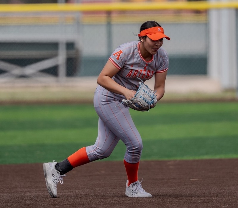 A teen girl in a grey and orange softball uniform leans forward as she prepares to take the ball out of her glove after making a catch.