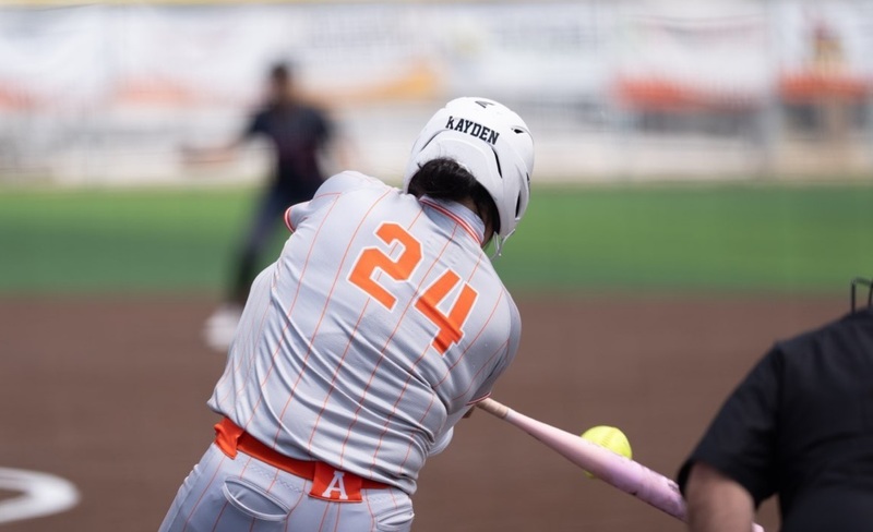 A teen girl in a grey and orange softball uniform is seen from behind as she puts the bat on the ball.