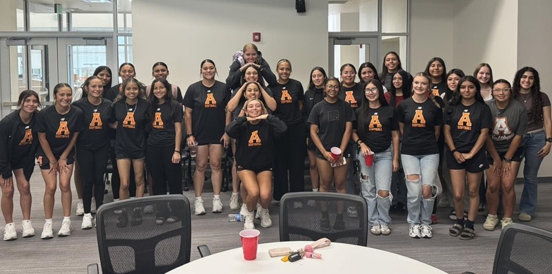 A high school softball team in black shirts with orange "As" on the front poses for a photo in a large room.