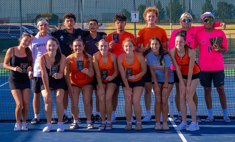Boys' and girls' tennis teams in orange and black uniforms pose for a photo on a tennis court. Many of the athletes hold small plaques.