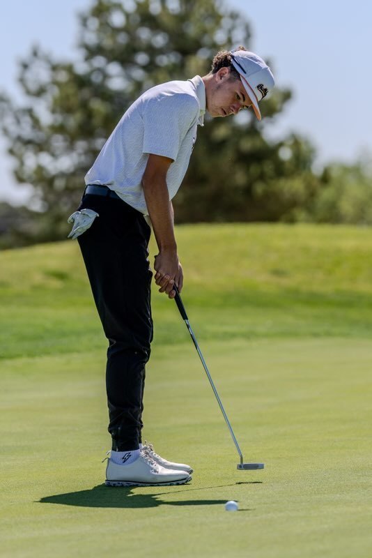 A teen boy in white and black watches a golf ball roll into a hole.