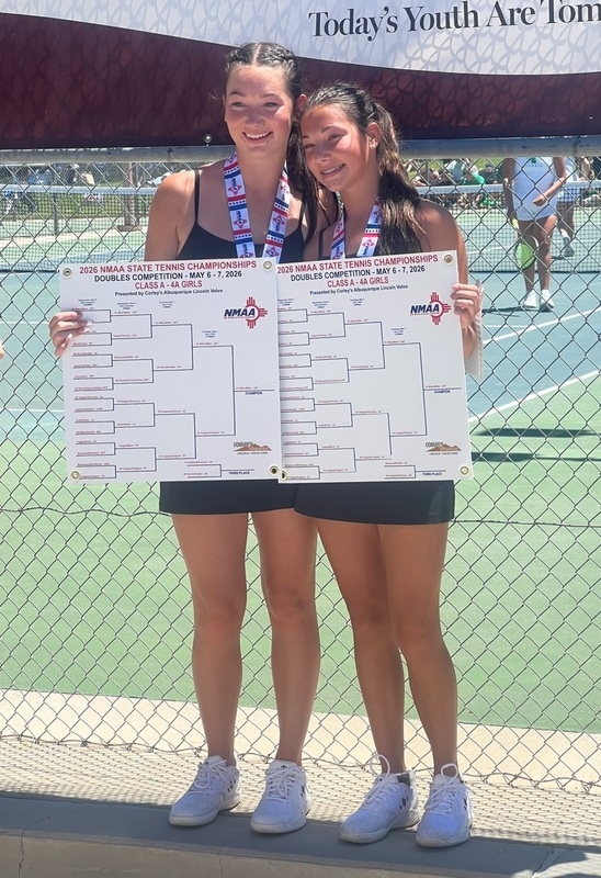Two teen girls pose for a photo in front of tennis courts, holding large bracket and wearing medals around their necks