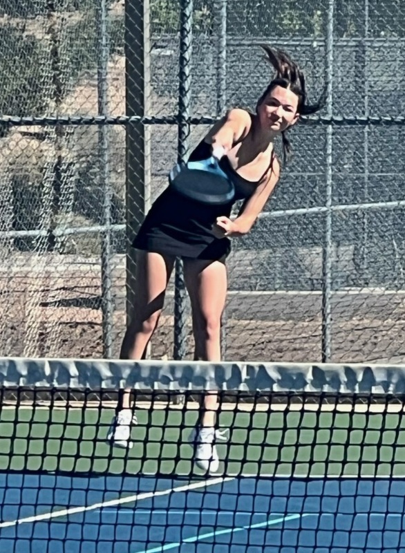 A teen girl in a black tennis uniform hops into the air as she powers her tennis racket forward.