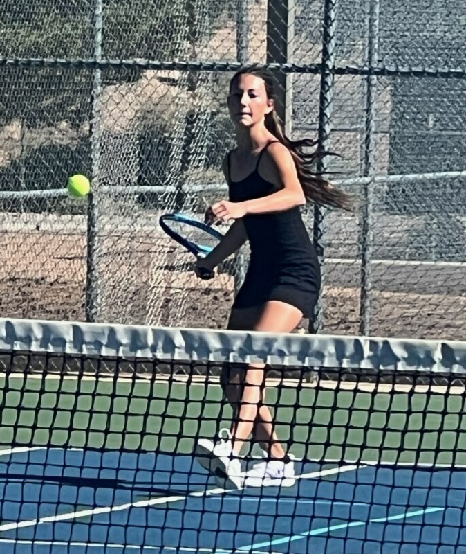 A teen girl in a black tennis uniform moves toward the net as she prepares to strike the ball.