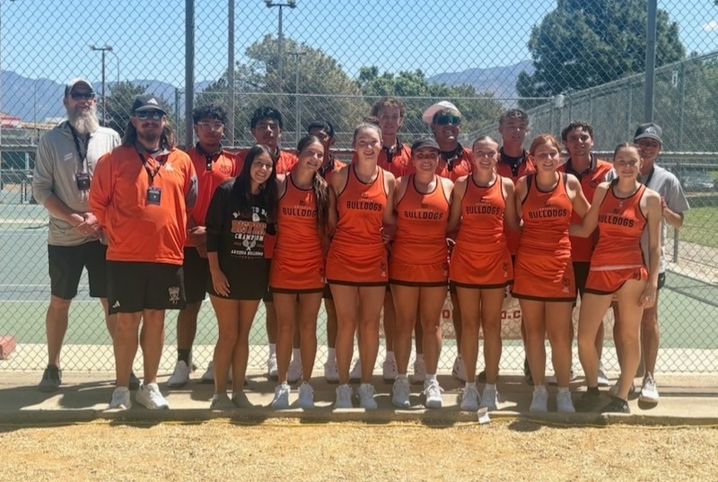 A boys' and a girls' tennis team in orange and black uniforms pose with a female coach and two male coaches in front of tennis courts.