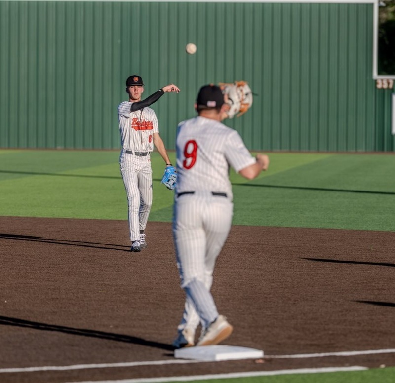 A teen boy in a black-and-white pin striped uniform with orange lettering tosses the ball to a teammate at first base.