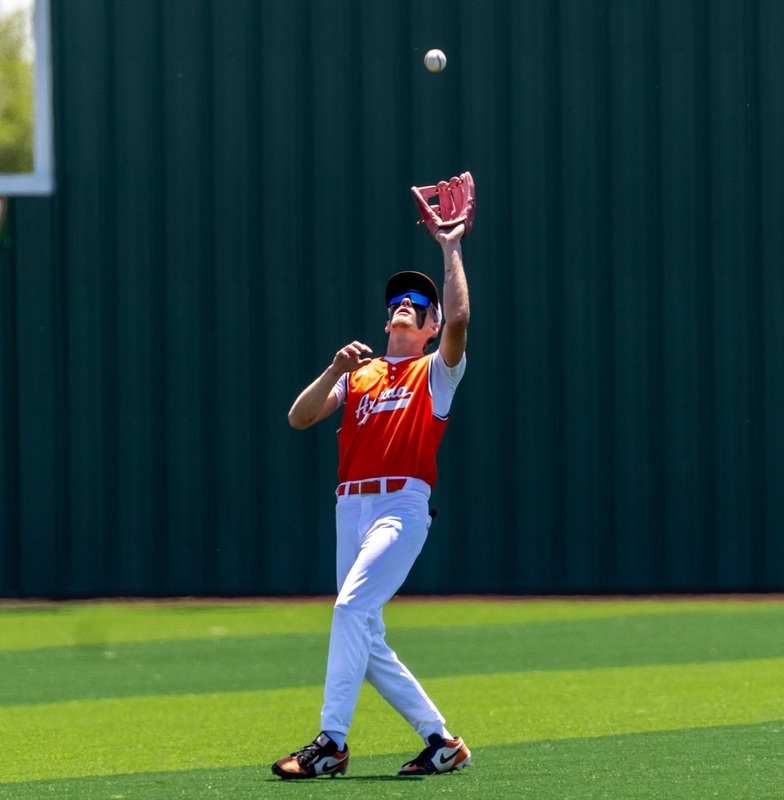 A teen boy in an orange and white baseball uniform and sunglasses holds his glove above his head with his left hand as he looks up at the approaching baseball.