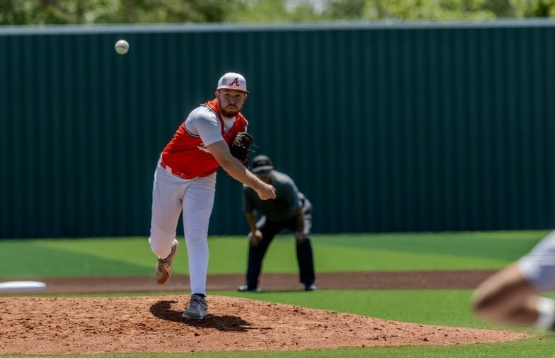 A teen boy in an orange and white baseball uniform throws the ball from the mound.
