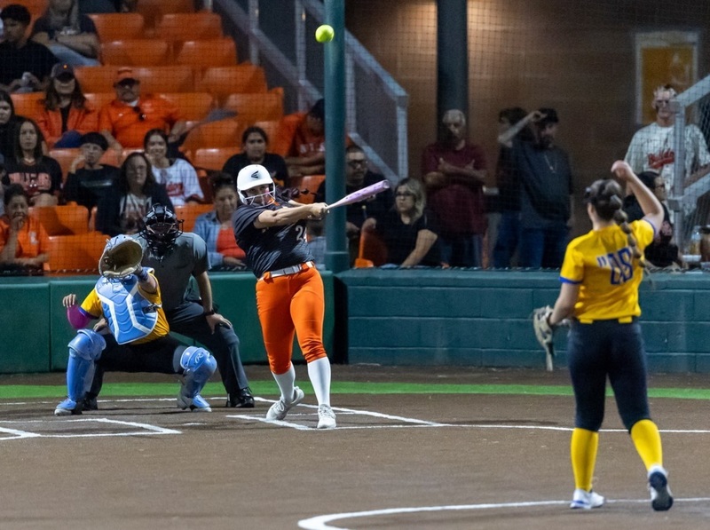 A teen girl in a black and orange softball uniform swings as the ball flies high off her bat. Also visible are the pitcher and catcher in yellow and black and an umpire.
