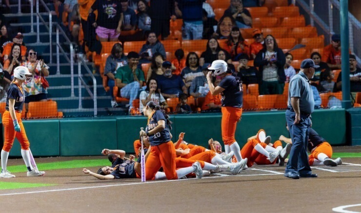 Teen girls in black and orange softball uniforms playfully fall to the ground as a teammate who just hit a home run arrives at the plate.