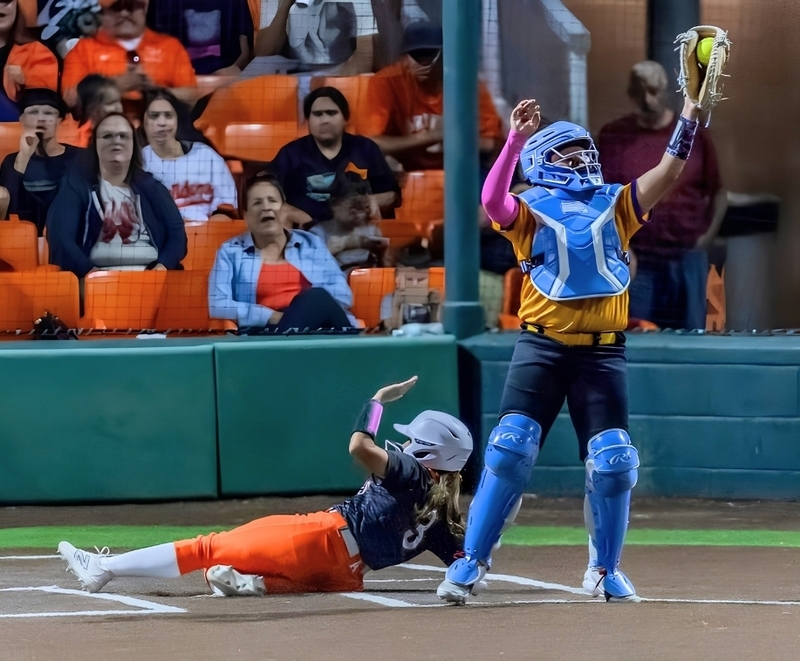 A teen girl in a black and orange softball uniform slides into home plate behind a catcher in a yellow and black uniform and blue equipment.