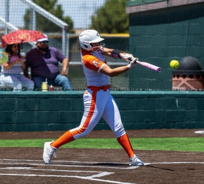 A teen girl in a white and orange softball uniform connects the bat with the ball.