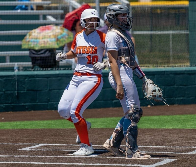 A teen girl in a white and orange softball uniform steps on home plate behind a catcher in white.
