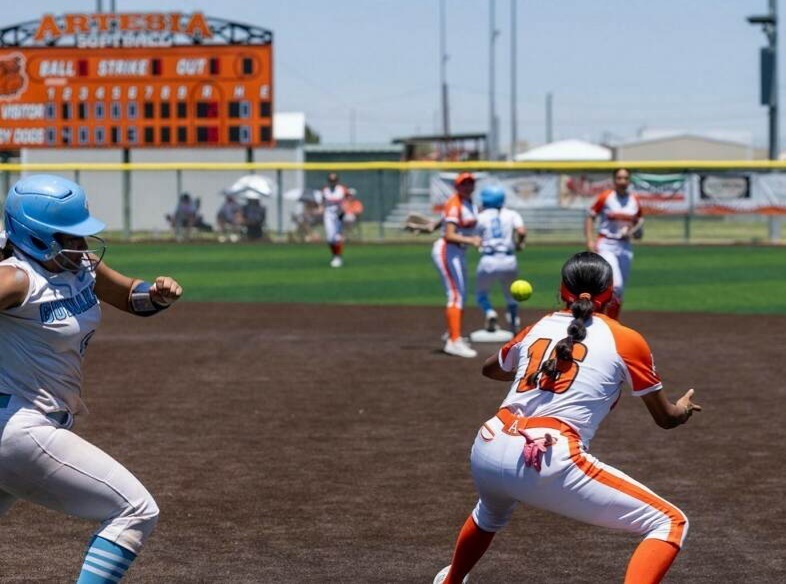 A teen girl in a white and orange softball uniform leans forward from first base as she prepares to catch the ball. A runner in white and blue can be seen headed toward her.