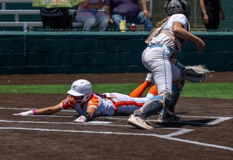 A teen girl in a white and orange softball uniform slides head first acfoss home plate as a catcher in white and blue looks for the ball.