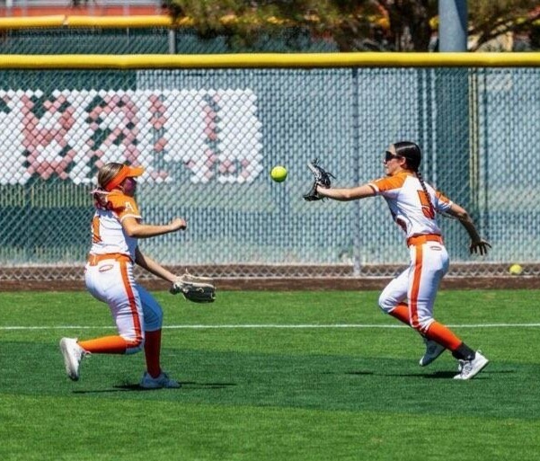 A teen girl in a white and orange softball uniform prepares to catch the ball in her glove in the outfield as a teammate looks on.