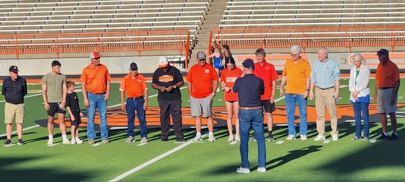 A row of men, along with a pair of young boys and one young girl, stand in a row on a football field.