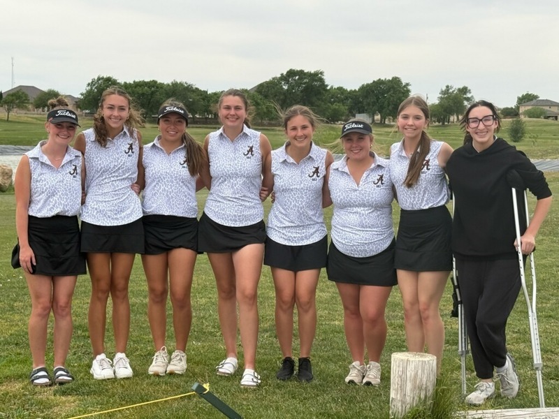 A group of teen girls in white and black golf uniforms poses for a photo on a course.
