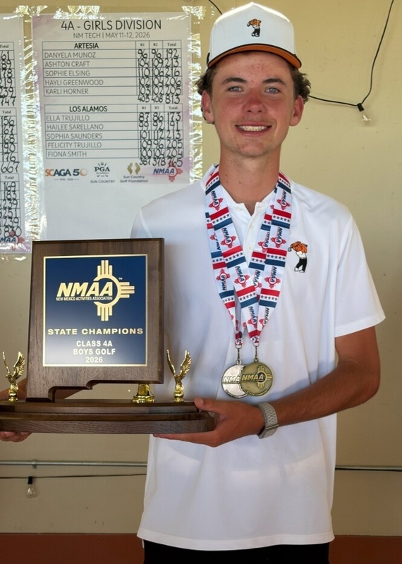 A teen boy in a white golf shirt and cap poses for a photo holding a blue trophy with two medals around his neck.