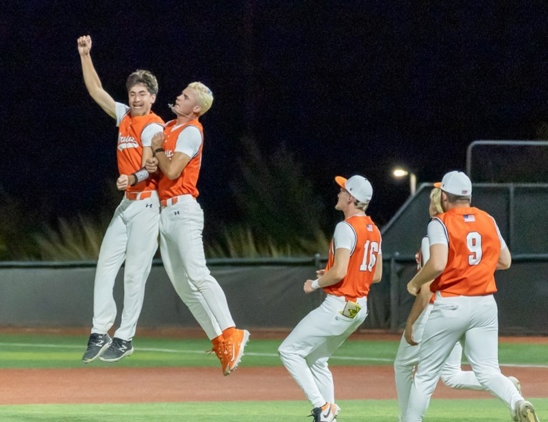Two teen boys in orange and white softball uniforms leap into the air with one another as three other boys run over to join them.