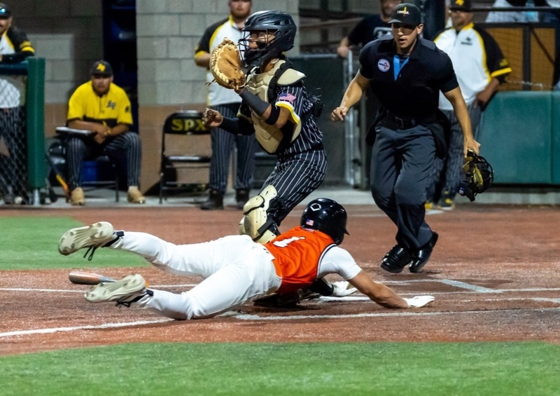 A teen boy in an orange and white baseball uniform slides head first across home plate as an opposing catcher in black and gold loos out toward the field and an umpire watches to ensure the runner has touched the plate.