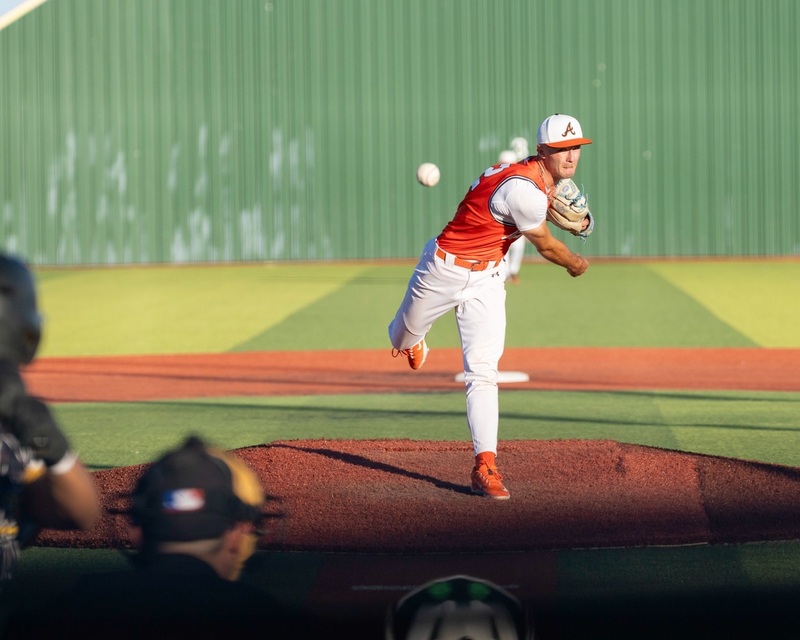 A teen boy in an orange and white baseball uniform fires a pitch from the mound.