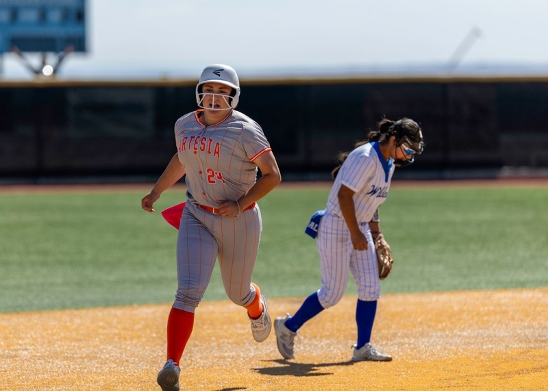 A teen girl in a grey and orange softball uniform runs between second and third as an opponent in white and blue looks dejected in the background.