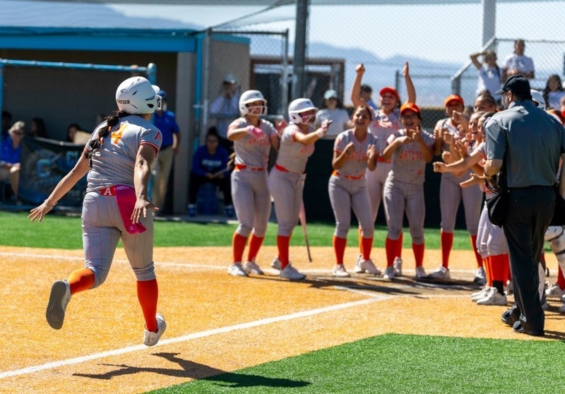 A teen girl in a grey and orange softball uniform runs toward home plate, where teammates are waiting to greet her.