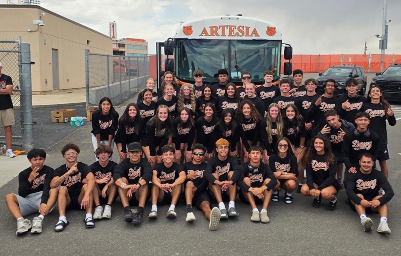 A large group of teen boys and girls in black t-shirts stand and sit in front of a bus in a parking lot.