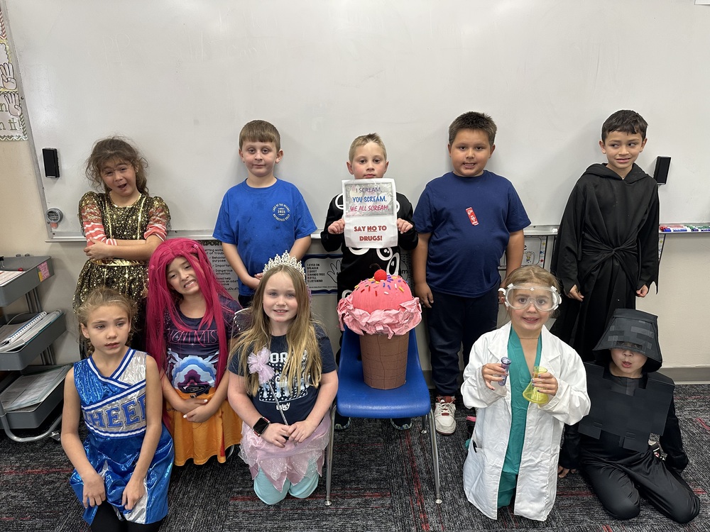 A group of elementary students pose in front of a whiteboard while wearing a variety of costumes, including a cheerleader, princesses, a scientist, and characters from different themes. In the center sits a large pink ice-cream-themed project with a sign that reads, “I scream, you scream, we all scream… SAY NO TO DRUGS!” The students smile proudly around the display.