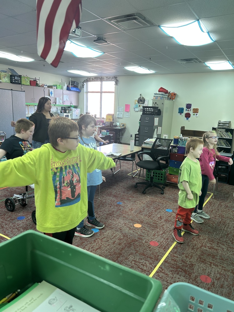 A group of elementary students stand spaced out in a classroom with their arms extended as they participate in a movement or exercise activity. A teacher stands with them demonstrating the motion. The classroom is bright, with colorful bins, posters, and supplies visible throughout the room.
