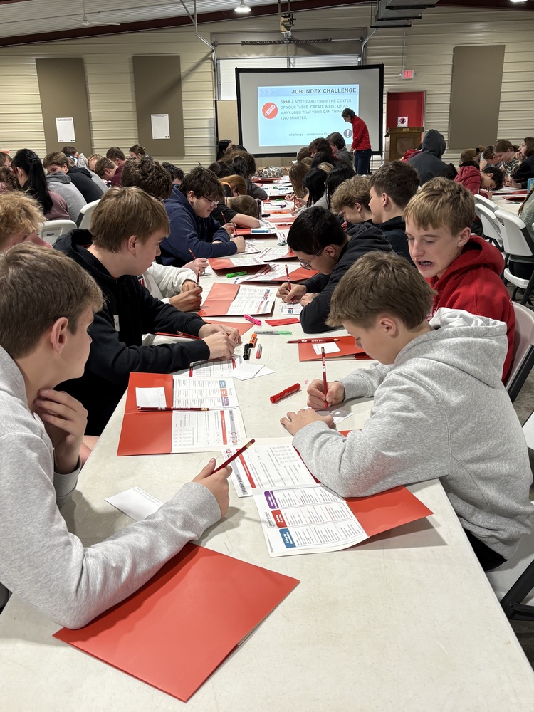 A large group of teenagers sit at long tables in a hall, writing on worksheets with pencils and red folders in front of them while participating in a classroom activity. At the front, an instructor stands near a projector screen displaying a "Job Index Challenge" prompt.
