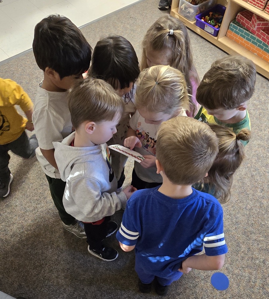 A small group of young children stand closely together in a classroom, looking down at and examining a small piece of paper or card while collaborating and sharing attention.