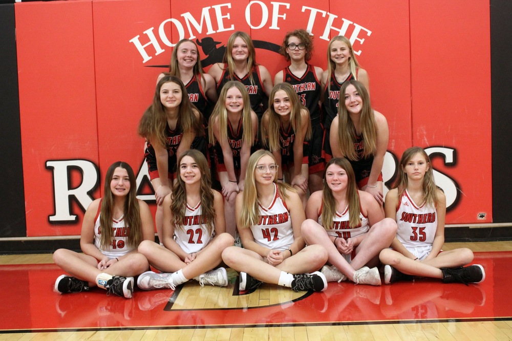 Junior high girls basketball team poses for a team photo in the gym, wearing Southern uniforms in red, black, and white. Players are arranged in rows in front of a red wall that reads “Home of the Raiders,” smiling toward the camera on the gym floor