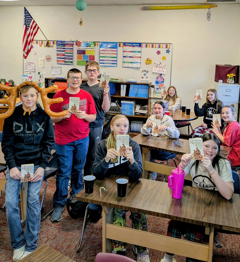 Group of middle school students gathered in a classroom, smiling and holding up copies of the same book during a class activity or celebration. Desks, bookshelves, posters, and classroom decorations are visible in the background, with students seated and standing together, showing excitement and pride in their reading.
