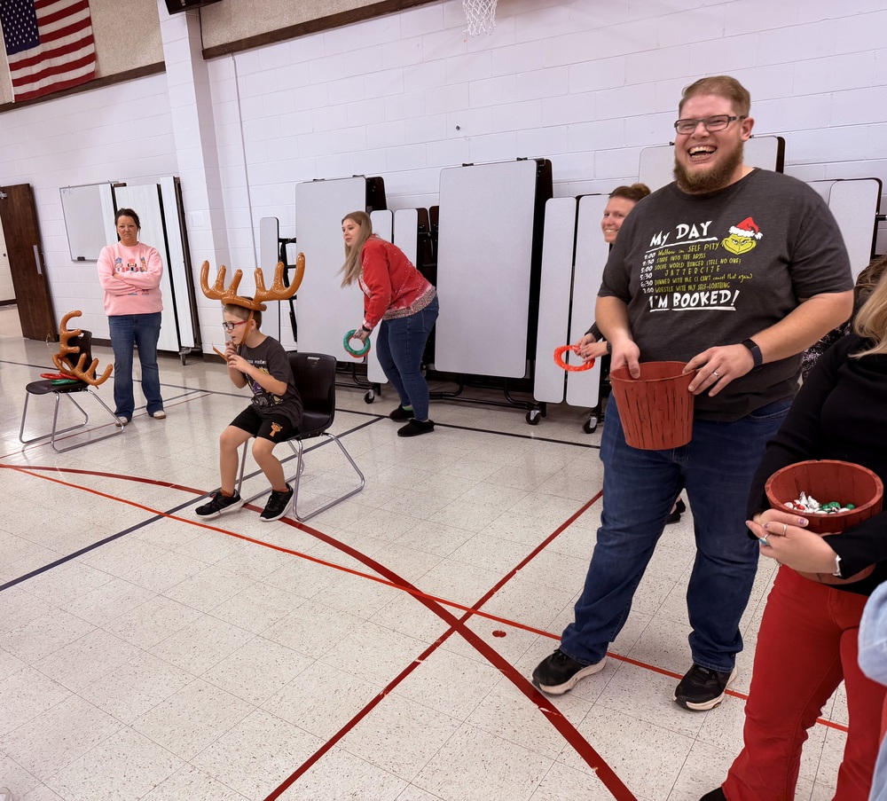 Elementary students and staff participate in a holiday-themed game in the school gym, with a child wearing reindeer antlers seated in a chair while adults laugh and toss rings into buckets during a festive Christmas activity.