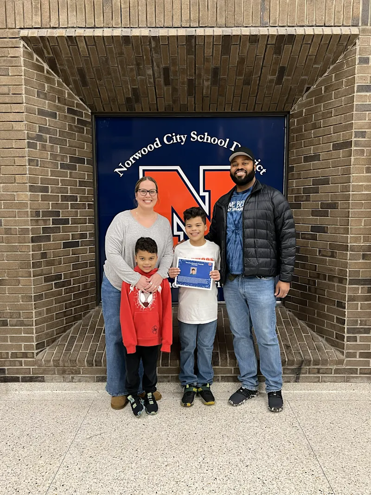A family of four poses for a photo in front of a brick alcove featuring a Norwood City School District sign with a large orange "N." A woman in a grey sweater and a man in a black puffer jacket stand behind two young boys. The boy in the center holds up a blue certificate, while the younger boy in front wears a red Spider-Man hoodie. All four are smiling at the camera.