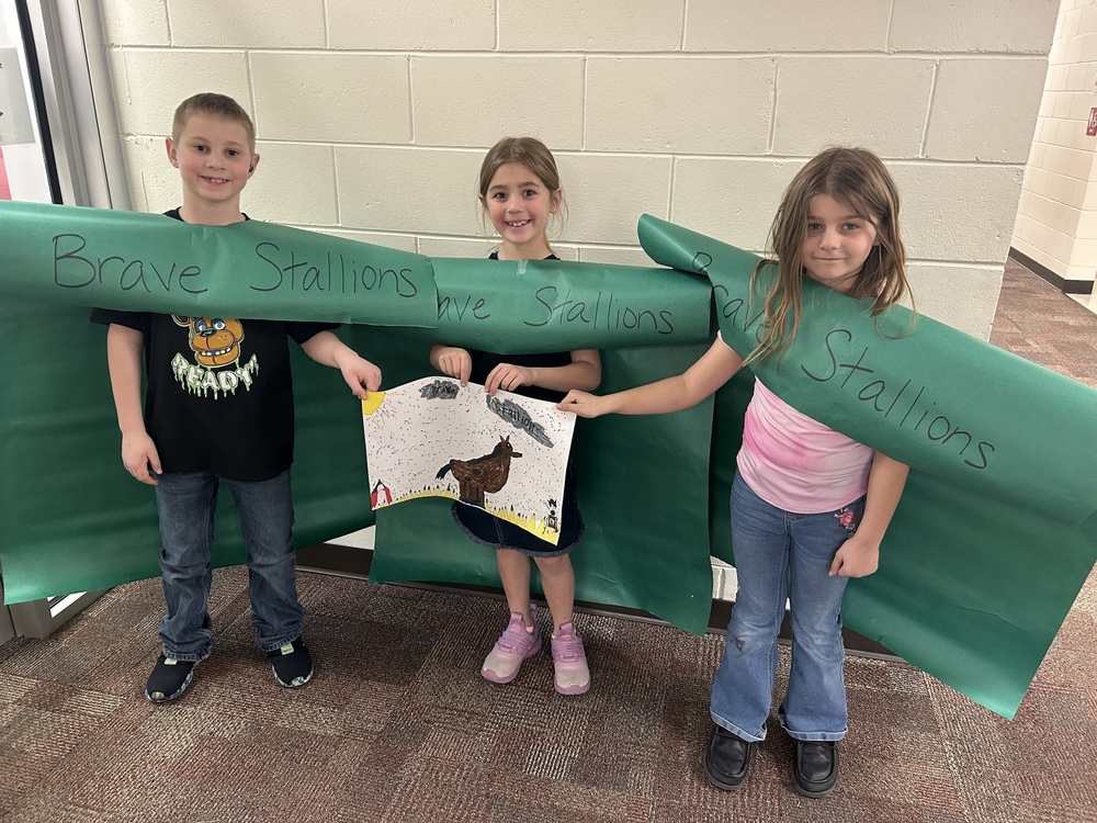 Three children stand smiling in front of a wall, holding a drawing of a horse. They hold a large green paper banner with "Brave Stallions" written on it. The atmosphere is cheerful.