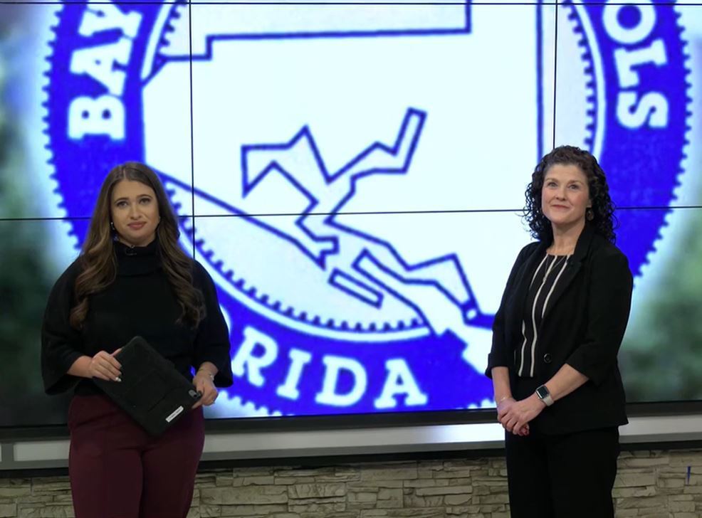 Two women standing in a studio setting in front of a large Bay District Schools logo displayed on a screen.